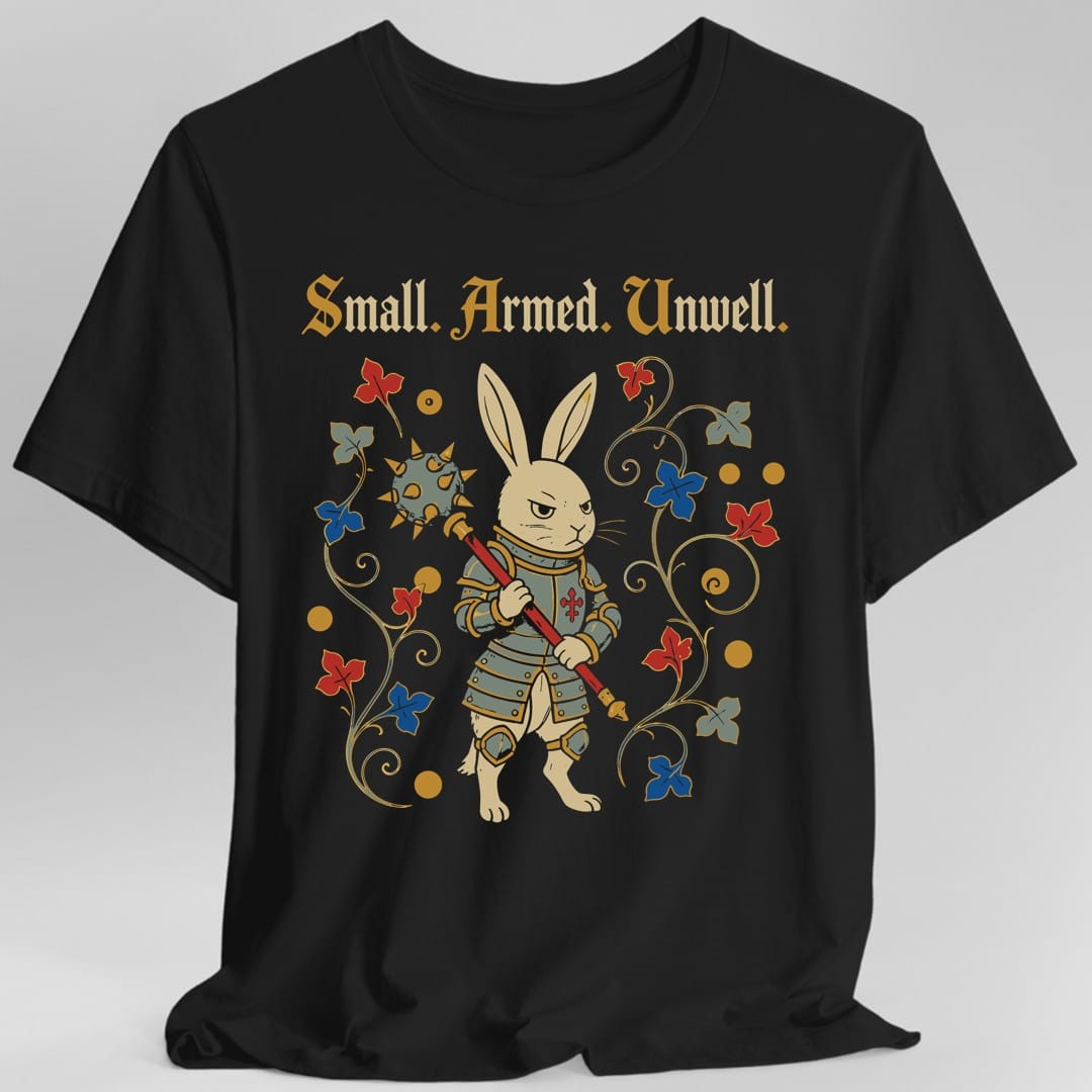 Black t-shirt. Reads "Small. Armed. Unwell." across the top in gold blackletter. Below, a rabbit stands upright in plate armour holding a long spear with a red cross on it, looking grimly ahead, framed by curling vines with red and blue flowers.