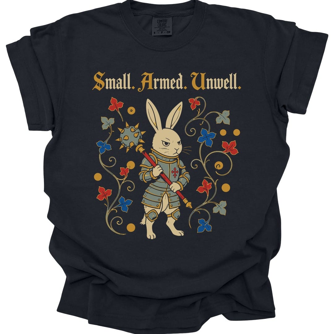 Black t-shirt. Reads "Small. Armed. Unwell." across the top in gold blackletter. Below, a rabbit stands upright in plate armor holding a long spear with a red cross on it, looking grimly ahead, framed by curling vines with red and blue flowers.