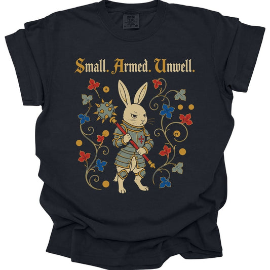 Black t-shirt. Reads "Small. Armed. Unwell." across the top in gold blackletter. Below, a rabbit stands upright in plate armor holding a long spear with a red cross on it, looking grimly ahead, framed by curling vines with red and blue flowers.
