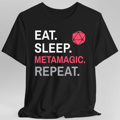 Black t-shirt. Four words stacked in heavy block capitals on four lines: "Eat." and "Sleep." in white, then "Metamagic." in hot pink, then "Repeat." in gray. A hot pink twenty-sided die sits in the top right corner beside the word Eat. All type, no illustration.