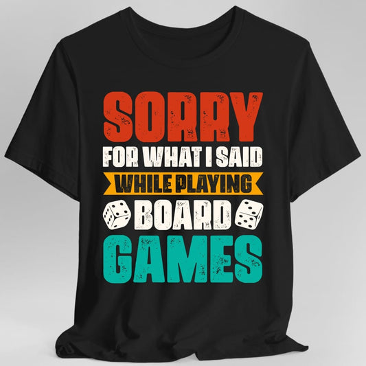 Black t-shirt. Reads "Sorry" in large red distressed capitals, "For what I said" in cream, "While playing" in a gold banner, "Board" and "Games" in teal, with a white die either side of the word Board. Retro distressed type throughout.