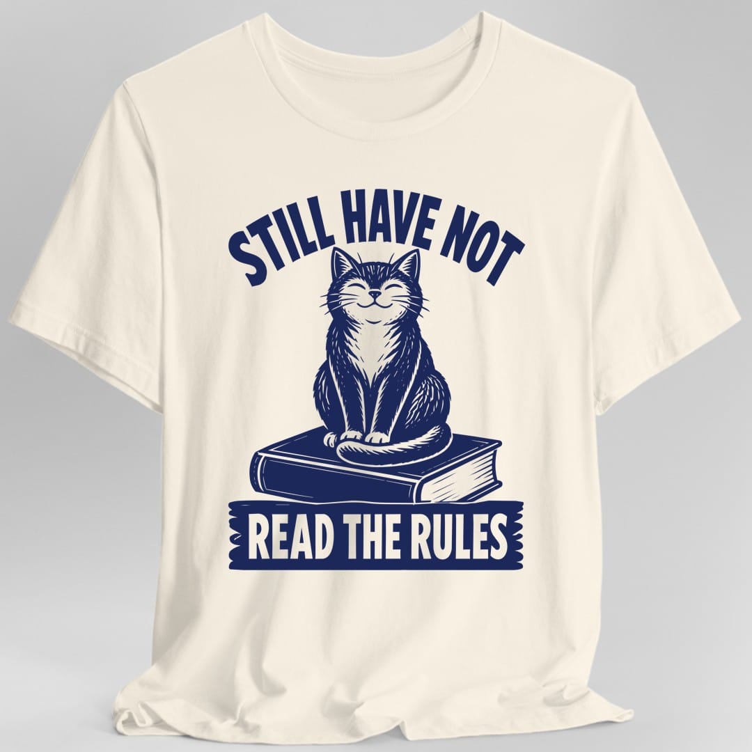 Natural t-shirt. The design reads STILL HAVE NOT in navy capitals arched across the top. Below it a contented cat with closed eyes and a slight smile sits upright on a closed hardback book, its tail curled around its front paws, drawn in navy woodcut hatching. Across the bottom a torn edged navy banner carries the words READ THE RULES in cut out letters that show the shirt color through them.