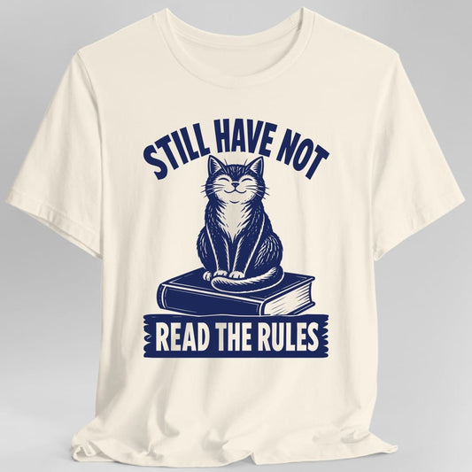 Natural t-shirt. The design reads STILL HAVE NOT in navy capitals arched across the top. Below it a contented cat with closed eyes and a slight smile sits upright on a closed hardback book, its tail curled around its front paws, drawn in navy woodcut hatching. Across the bottom a torn edged navy banner carries the words READ THE RULES in cut out letters that show the shirt color through them.