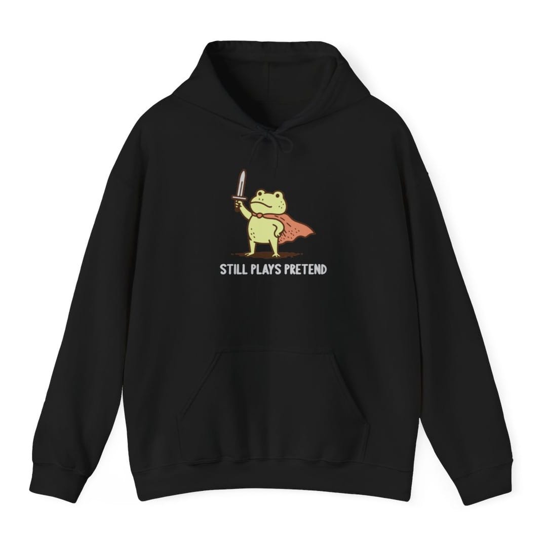 Black hoodie. A small green frog stands upright in a coral cape, holding a sword up in one hand, its shadow beneath it. Underneath, "Still plays pretend" in cream capitals. Simple soft illustration.