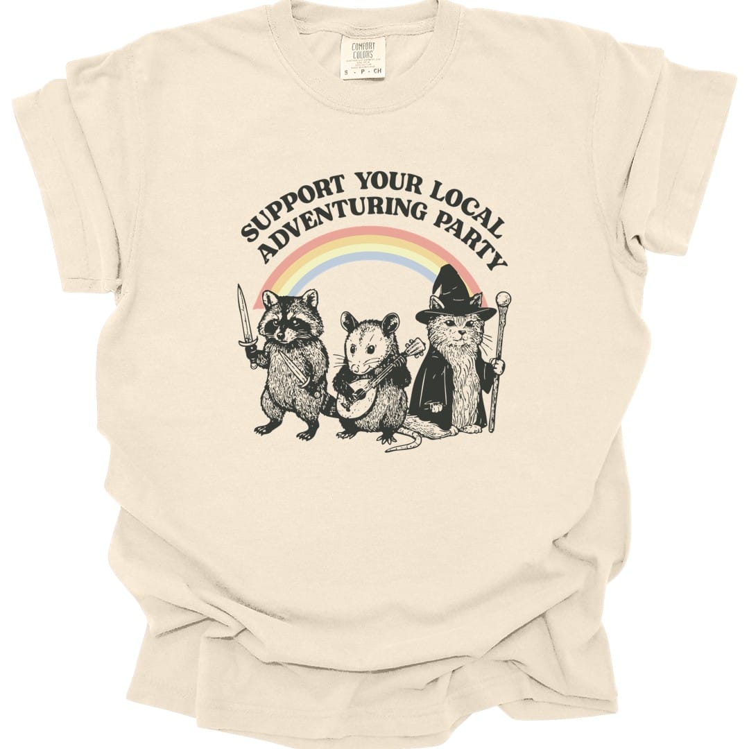 Ivory t-shirt. Reads "Support your local adventuring party" curving across the top in dark capitals. Below, three animals stand in a row: a raccoon holding a sword, an opossum with a dagger, and a cat in a pointed wizard hat with a staff. A soft pastel rainbow arcs behind them. Engraved linework over flat pastel color.