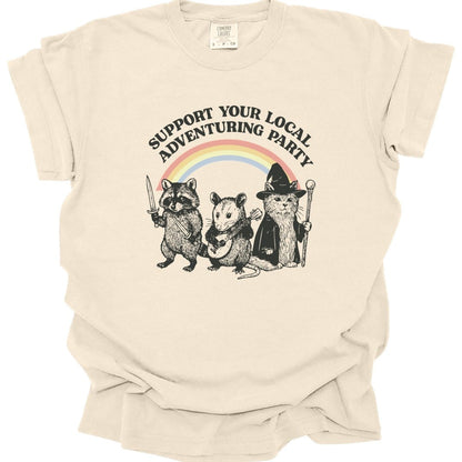 Ivory t-shirt. Reads "Support your local adventuring party" curving across the top in dark capitals. Below, three animals stand in a row: a raccoon holding a sword, an opossum with a dagger, and a cat in a pointed wizard hat with a staff. A soft pastel rainbow arcs behind them. Engraved linework over flat pastel color.