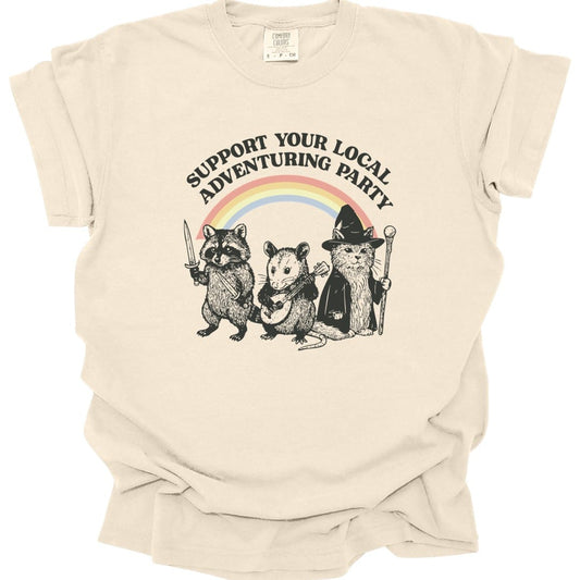 Ivory t-shirt. Reads "Support your local adventuring party" curving across the top in dark capitals. Below, three animals stand in a row: a raccoon holding a sword, an opossum with a dagger, and a cat in a pointed wizard hat with a staff. A soft pastel rainbow arcs behind them. Engraved linework over flat pastel color.