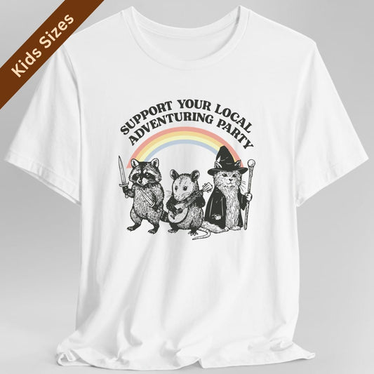White kids t-shirt. Reads "Support your local adventuring party" curving across the top in dark capitals. Below, three animals stand in a row: a raccoon holding a sword, an opossum with a dagger, and a cat in a pointed wizard hat with a staff. A soft pastel rainbow arcs behind them. Engraved linework over flat pastel color.