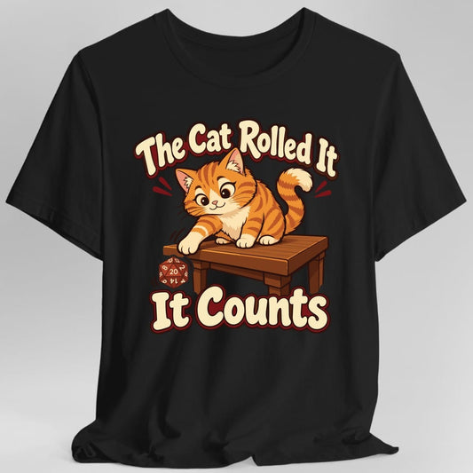 Black t-shirt. Reads "The Cat Rolled It" in a curve across the top and "It Counts" across the bottom, in rounded cream letters outlined in dark red. Between them a plump orange tabby kitten crouches on a wooden table, one paw stretched out, having just batted a red twenty sided die off the edge. The die is mid-tumble and shows a 20. Soft cartoon illustration in orange, cream and warm brown.