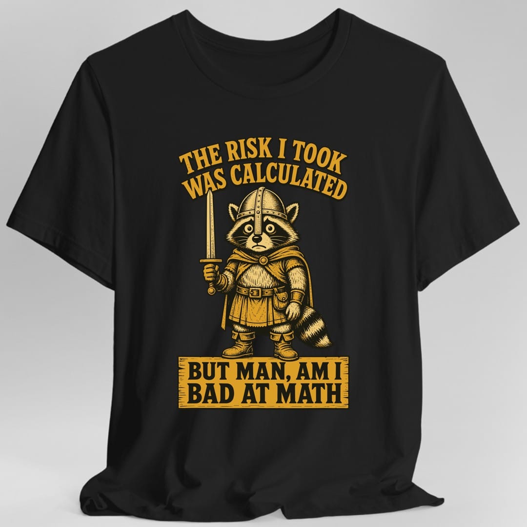 Black t-shirt. The design reads The Risk I Took Was Calculated in arched gold capitals across the top, and But Man, Am I Bad At Math in black capitals on a gold banner across the bottom. Between them a worried raccoon stands in a domed helmet, hooded cape, belted tunic and boots, holding a sword upright in one paw, its ringed tail curling behind. Detailed gold and black engraving style.