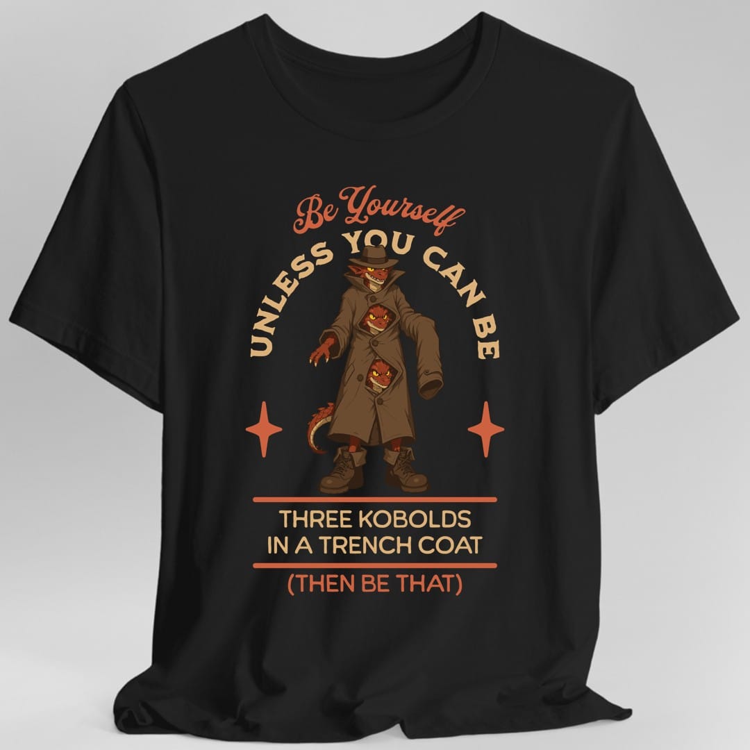 Black t-shirt. The design reads "Be yourself" in script at the top, then "unless you can be" curving around the picture, then "Three kobolds in a trench coat" on two lines below, then "(then be that)" at the bottom. In the middle, three small red kobolds are stacked inside one long tan trench coat and a fedora, the top one grinning out from under the hat brim and the other two peering through gaps between the buttons, with a tail poking out of the hem. Bold cartoon art in tan, rust and dark red.