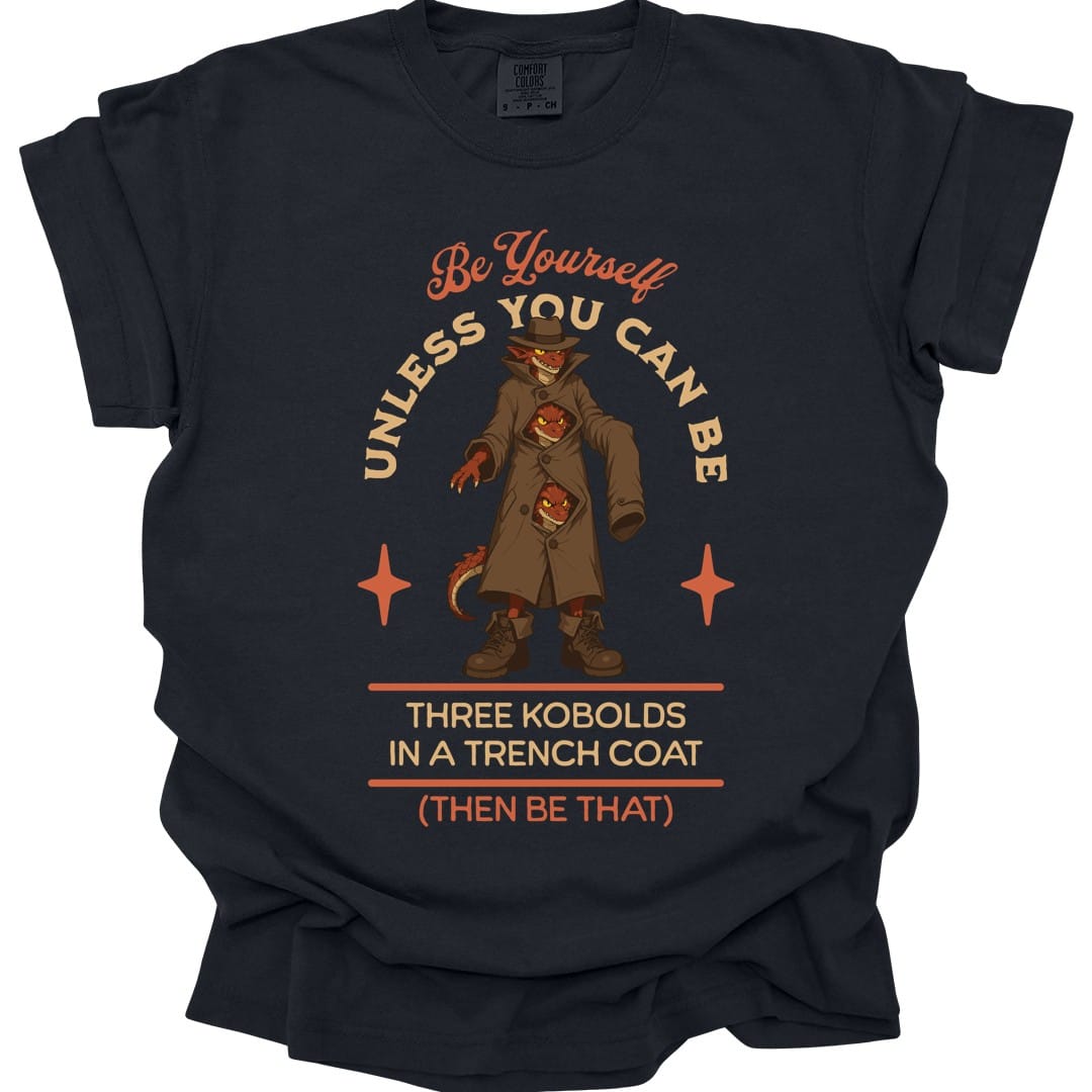 Black t-shirt. The design reads "Be yourself" in script at the top, then "unless you can be" curving around the picture, then "Three kobolds in a trench coat" on two lines below, then "(then be that)" at the bottom. In the middle, three small red kobolds are stacked inside one long tan trench coat and a fedora, the top one grinning out from under the hat brim and the other two peering through gaps between the buttons, with a tail poking out of the hem. Bold cartoon art in tan, rust and dark red.