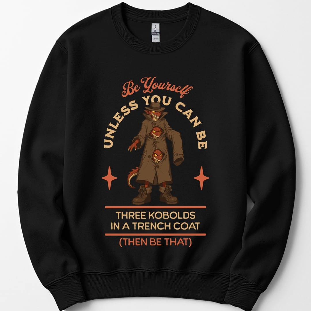 Black sweatshirt. The design reads "Be yourself" in script at the top, then "unless you can be" curving around the picture, then "Three kobolds in a trench coat" on two lines below, then "(then be that)" at the bottom. In the middle, three small red kobolds are stacked inside one long tan trench coat and a fedora, the top one grinning out from under the hat brim and the other two peering through gaps between the buttons, with a tail poking out of the hem. Bold cartoon art in tan, rust and dark red.