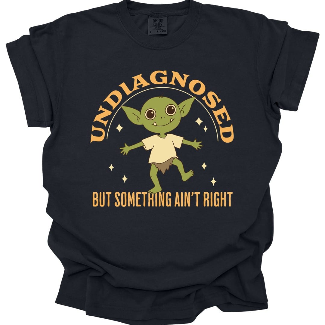 Black t-shirt. Reads "Undiagnosed" curving across the top and "But something ain't right" across the bottom in gold capitals. Between them a small green goblin stands with arms spread wide and a wide grin, wearing a simple cream tunic, sparkles either side.