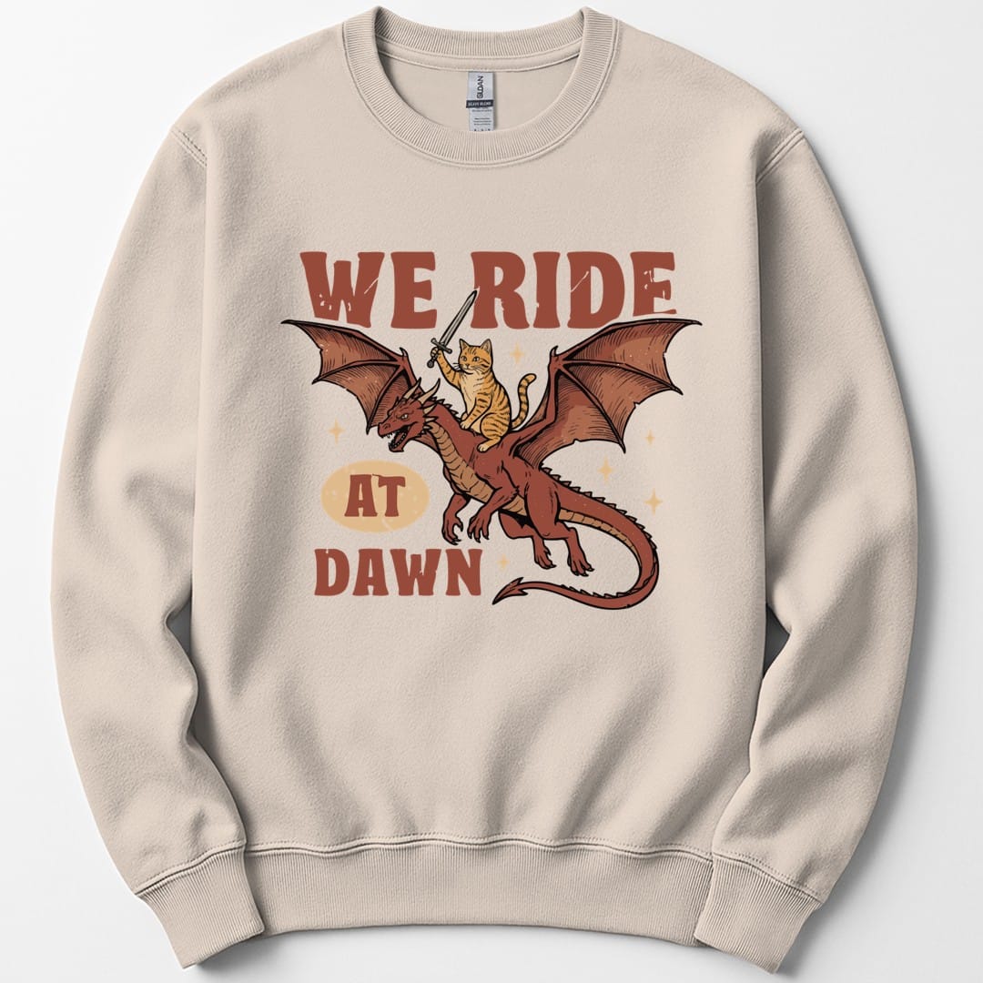 Sand sweatshirt. Reads "We ride" across the top in red capitals and "At dawn" across the bottom, with "at dawn" partly inside a cream oval. Between them a ginger tabby cat rides on the back of a red dragon in flight, holding a tiny sword aloft. Retro cartoon style.