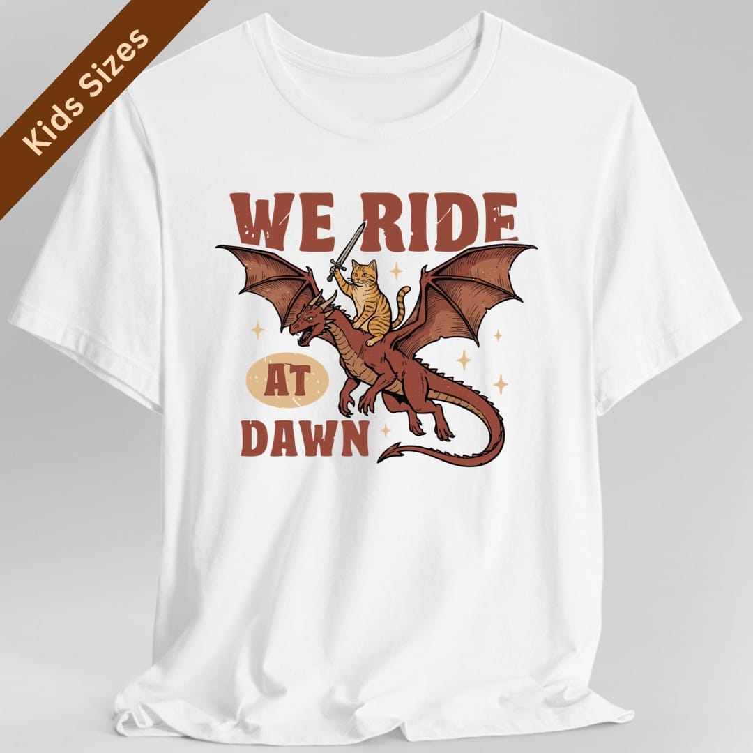 White kids t-shirt. Reads "We ride" across the top in red capitals and "At dawn" across the bottom, with "at dawn" partly inside a cream oval. Between them a ginger tabby cat rides on the back of a red dragon in flight, holding a tiny sword aloft. Retro cartoon style.