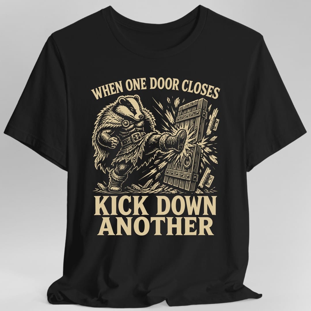Black t-shirt. The design reads "WHEN ONE DOOR CLOSES" curved across the top and "KICK DOWN ANOTHER" in two big lines at the bottom. In the middle, a badger in a studded belt and boots swings a boot into a heavy plank door, knocking it off its hinges in a burst of flying splinters. All of it is drawn in one warm sand color in a scratchy woodcut style.