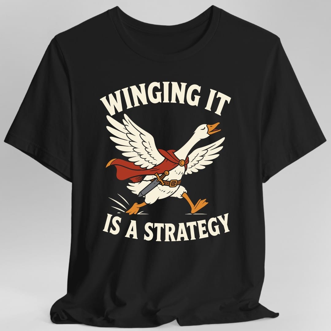 Black t-shirt. The design reads "WINGING IT" arched above and "IS A STRATEGY" curved below, in cream letters. Between them a cream goose charges forward with both wings spread, wearing a red cape clasped at the throat and a brown belt with a sword hanging from it. Its orange beak is open and its orange feet are mid-run, with small motion lines behind.
