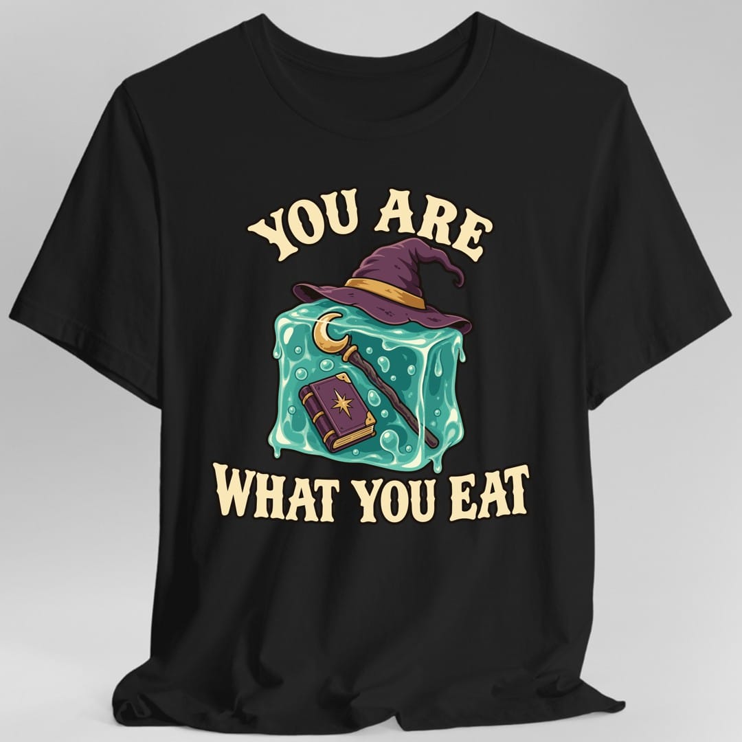 Black t-shirt. The design reads "You Are What You Eat", in chunky cream lettering arched above and below the art. In the middle is a teal gelatinous cube with a bubbled, dripping surface, wearing a purple wizard hat with a gold band. Floating inside the cube are a purple spellbook with a gold star on its cover and a long wooden staff topped with a gold crescent moon. Bold outlined cartoon art in teal, purple, gold and cream.